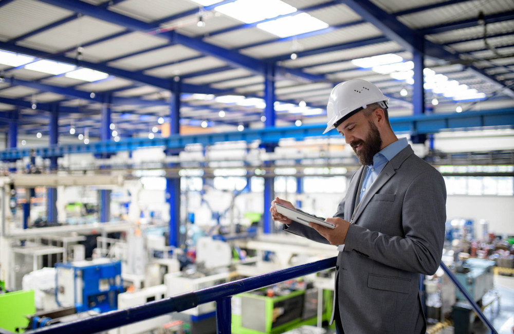 Ein Mann im Anzug übersieht eine Produktionshalle, trägt einen Schutzhelm und liest etwas auf einem Tablet.