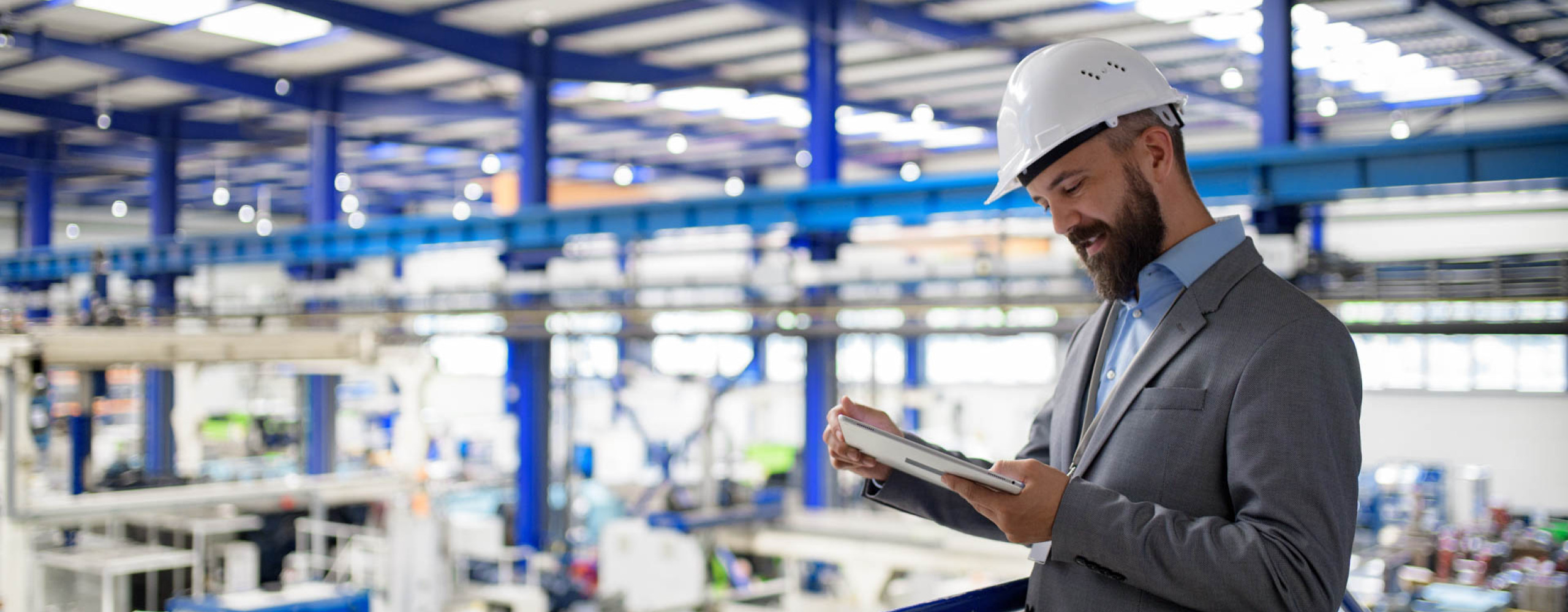 Ein Mann im Anzug übersieht eine Produktionshalle, trägt einen Schutzhelm und liest etwas auf einem Tablet.
