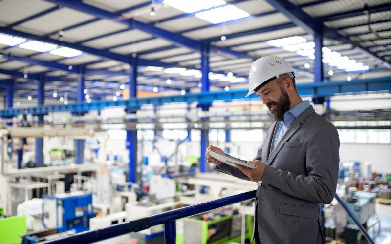 Ein Mann im Anzug übersieht eine Produktionshalle, trägt einen Schutzhelm und liest etwas auf einem Tablet.