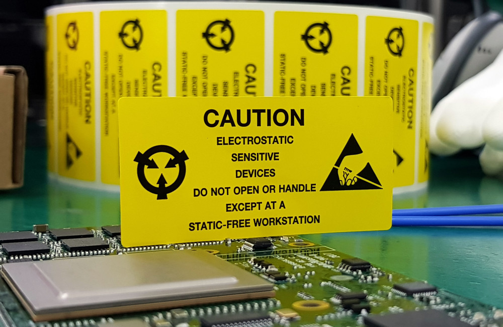 Eine elektronische Platine liegt auf einem ESD-Arbeitstisch. Ein gelbes Schild weist auf die Risiken von elektrostatischer Entladung hin.
