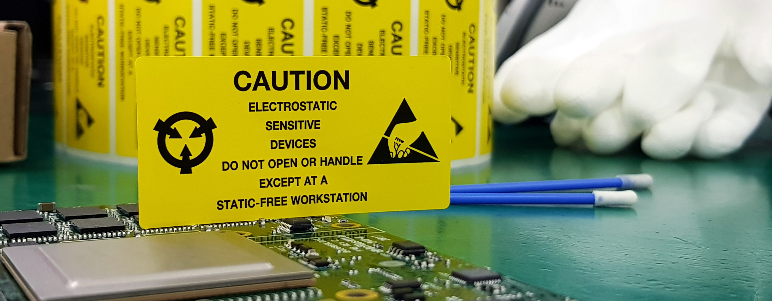Eine elektronische Platine liegt auf einem ESD-Arbeitstisch. Ein gelbes Schild weist auf die Risiken von elektrostatischer Entladung hin.