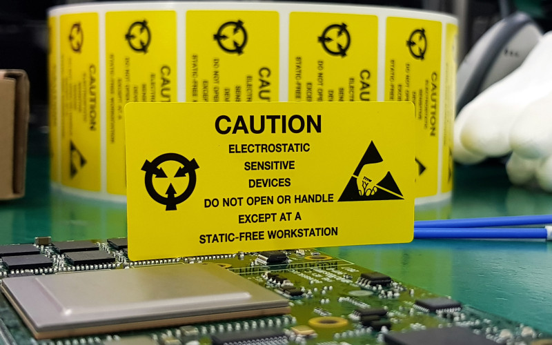 Eine elektronische Platine liegt auf einem ESD-Arbeitstisch. Ein gelbes Schild weist auf die Risiken von elektrostatischer Entladung hin.