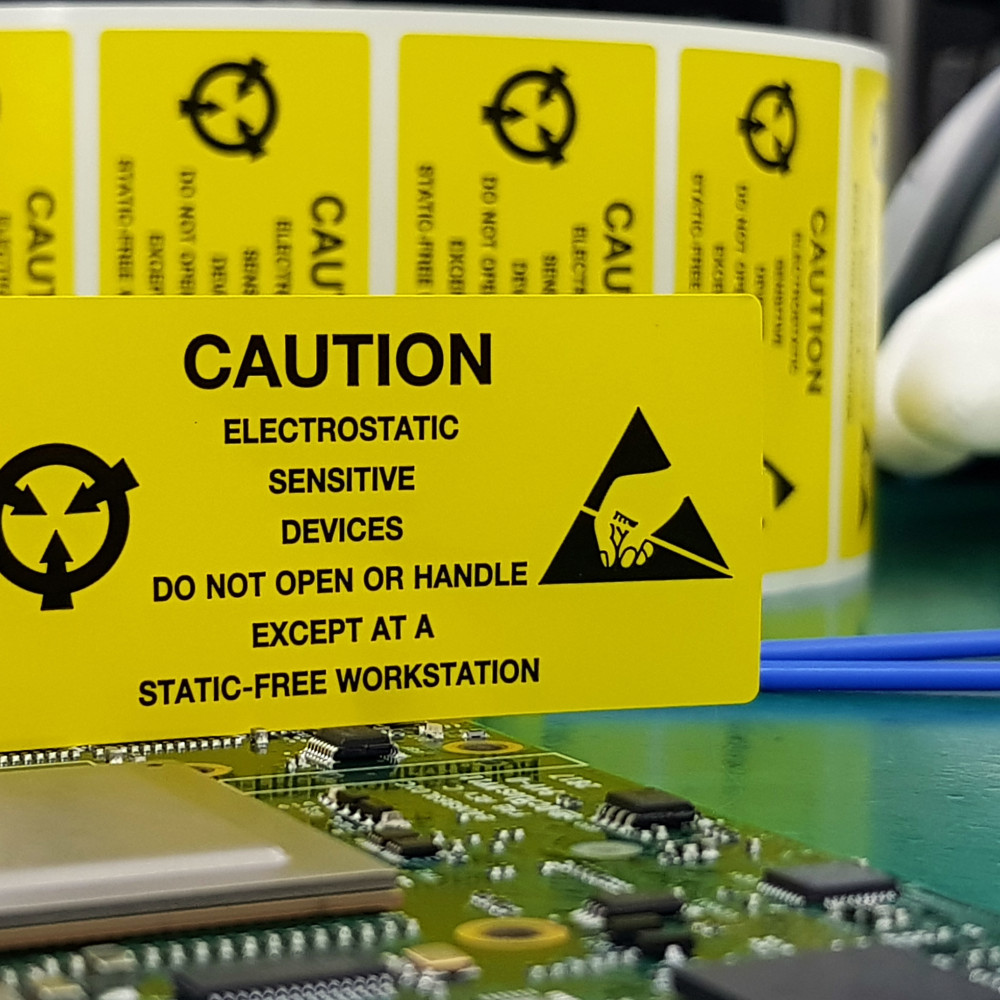 Eine elektronische Platine liegt auf einem ESD-Arbeitstisch. Ein gelbes Schild weist auf die Risiken von elektrostatischer Entladung hin.