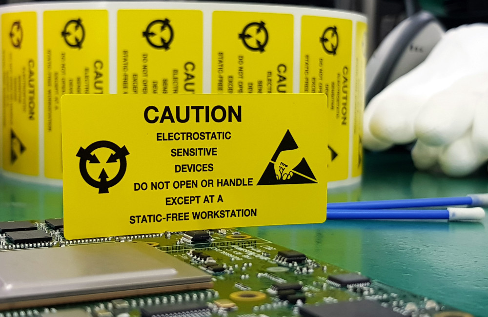 Eine elektronische Platine liegt auf einem ESD-Arbeitstisch. Ein gelbes Schild weist auf die Risiken von elektrostatischer Entladung hin.