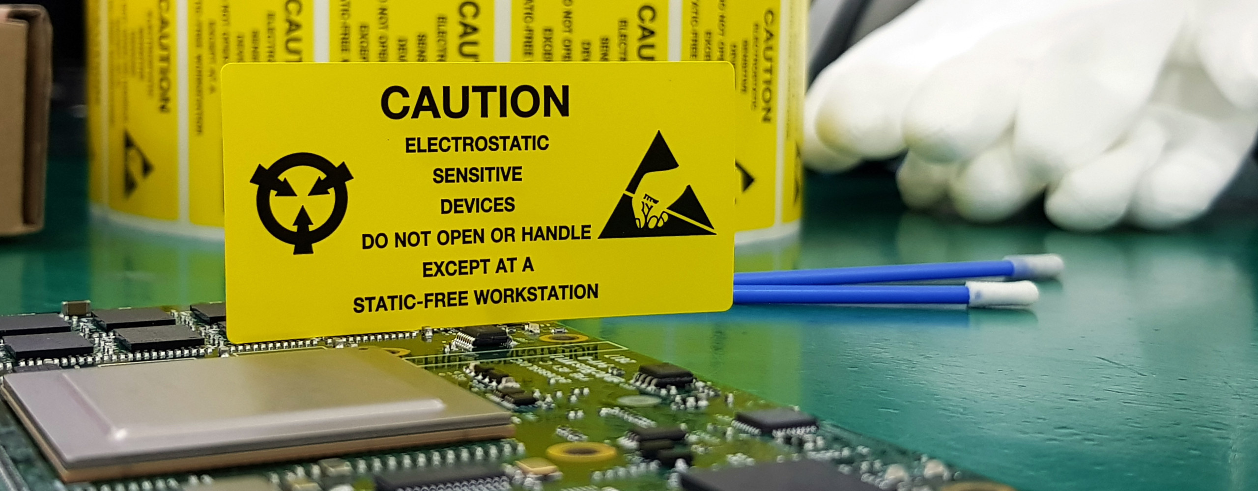 Eine elektronische Platine liegt auf einem ESD-Arbeitstisch. Ein gelbes Schild weist auf die Risiken von elektrostatischer Entladung hin.