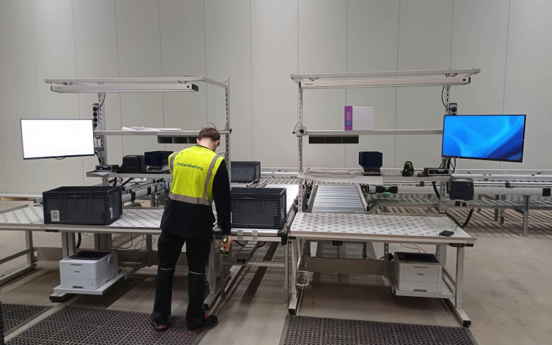 Ein Mann mit Warnweste befindet sich in einer Produktionshalle und arbeitet an einer Logistik-Packstation.