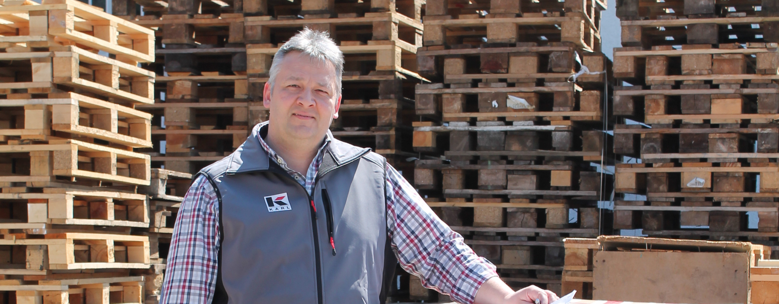 Mann steht neben einem großen Karton auf einer Palette, umgeben von gestapelten Holzpaletten in einem Lagerbereich im Freien.